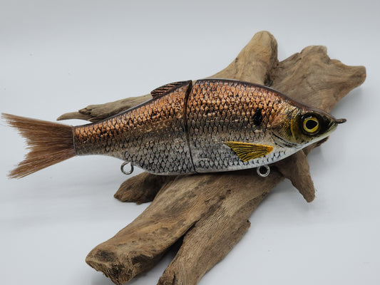 A realistic looking glidebait fishing lure with mottled brown and silver colors, positioned on a wooden surface.