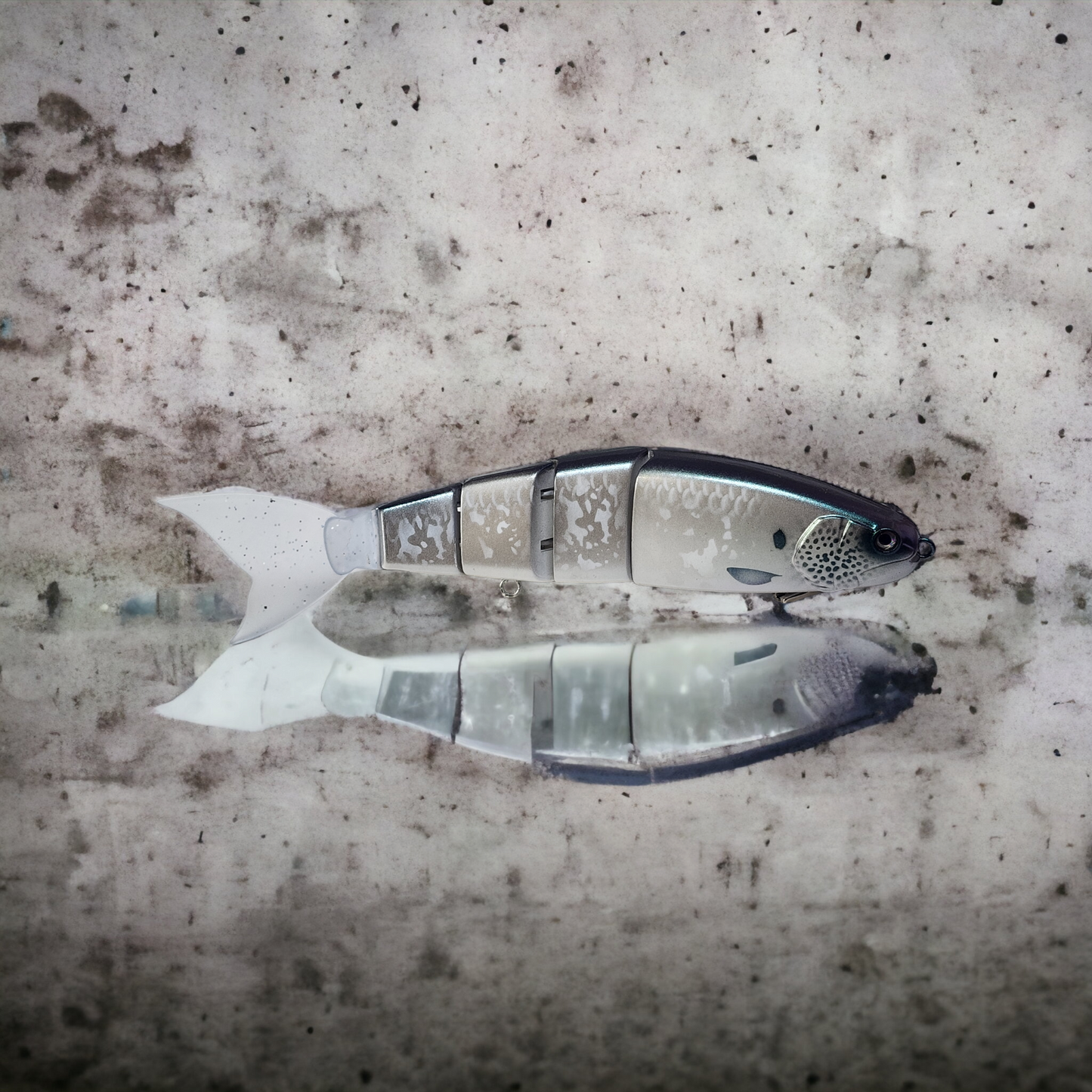 A swimbait lure with a silver head and tail, and a translucent body with black and white patterns, lying on a textured grey surface.